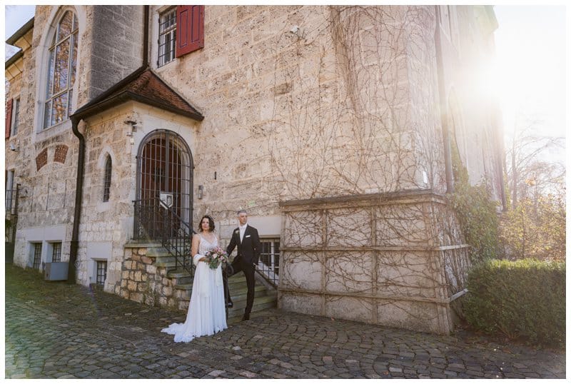 Brautpaarportrait von weiter weg an einem Schloss. Er hält sich am Geländer und hat einen Fuß auf den Treppenstufen